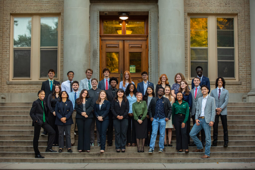 A large, diverse group of young adults dressed in business professional attire posing in rows on the wide stone steps of a brick building with large columns and tall wooden doors.